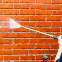Osoba w czapce i okularach z pomarańczową osłoną myje czerwoną ceglaną ścianę myjką ciśnieniową.